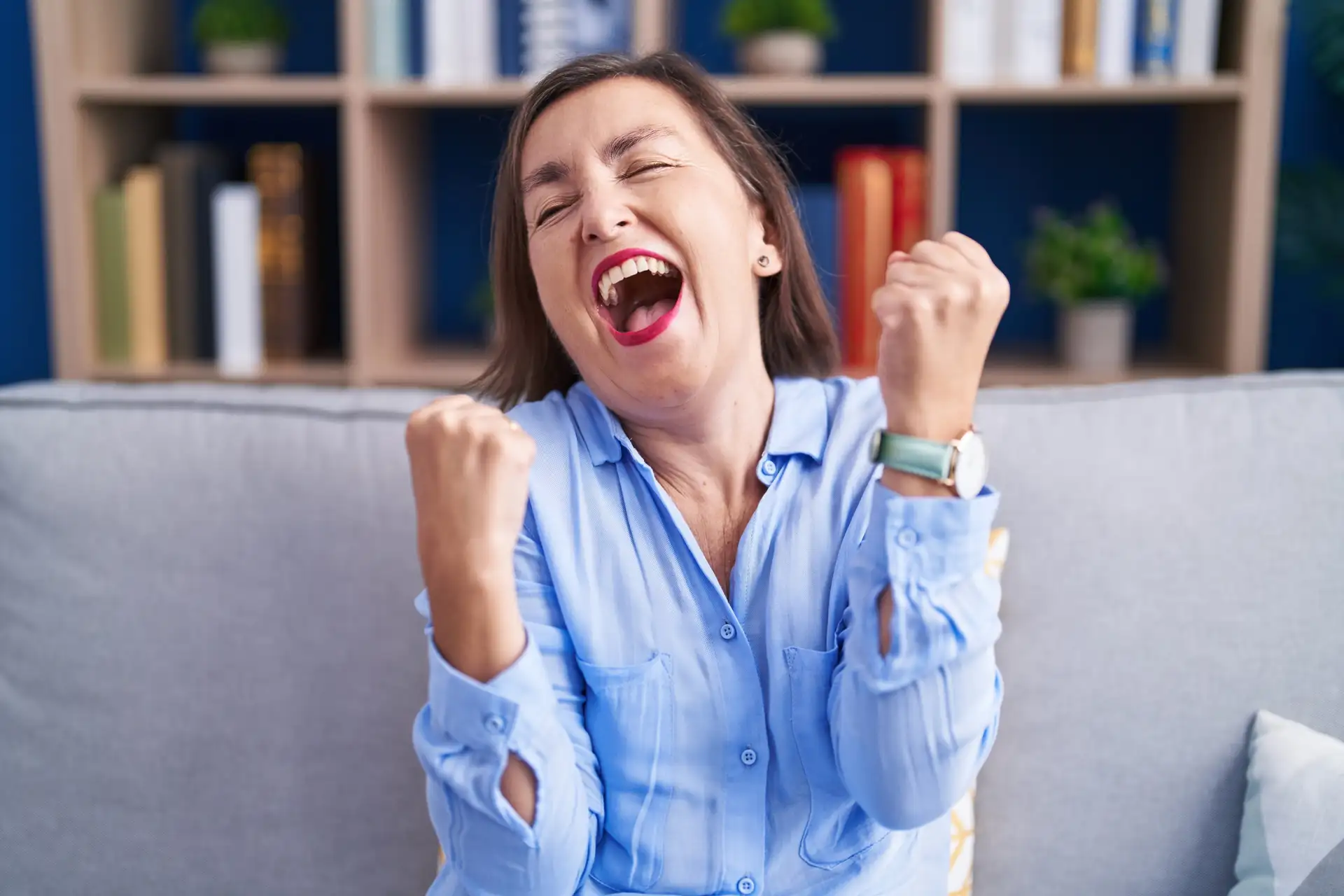 Middle age hispanic woman sitting on the sofa at home very happy and excited doing winner gesture with arms raised, smiling and screaming for success. celebration concept