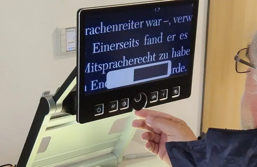 Zwei Personen testen gemeinsam ein Bildschirmlesegerät in einer Bibliothek. Das Gerät dient zur Vergrößerung von Texten für Menschen mit Sehbeeinträchtigung.