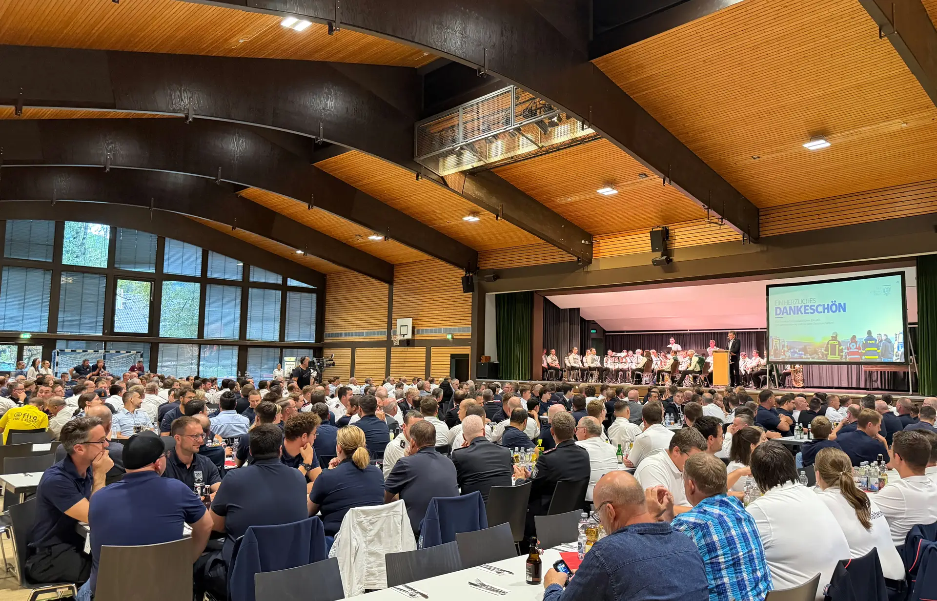 In einer vollen Halle sind Zuschauer bei einer Veranstaltung zu sehen; im Vordergrund stehen Personen.
